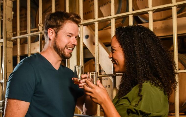 man and woman holding whiskey glasses next to whiskey barrels